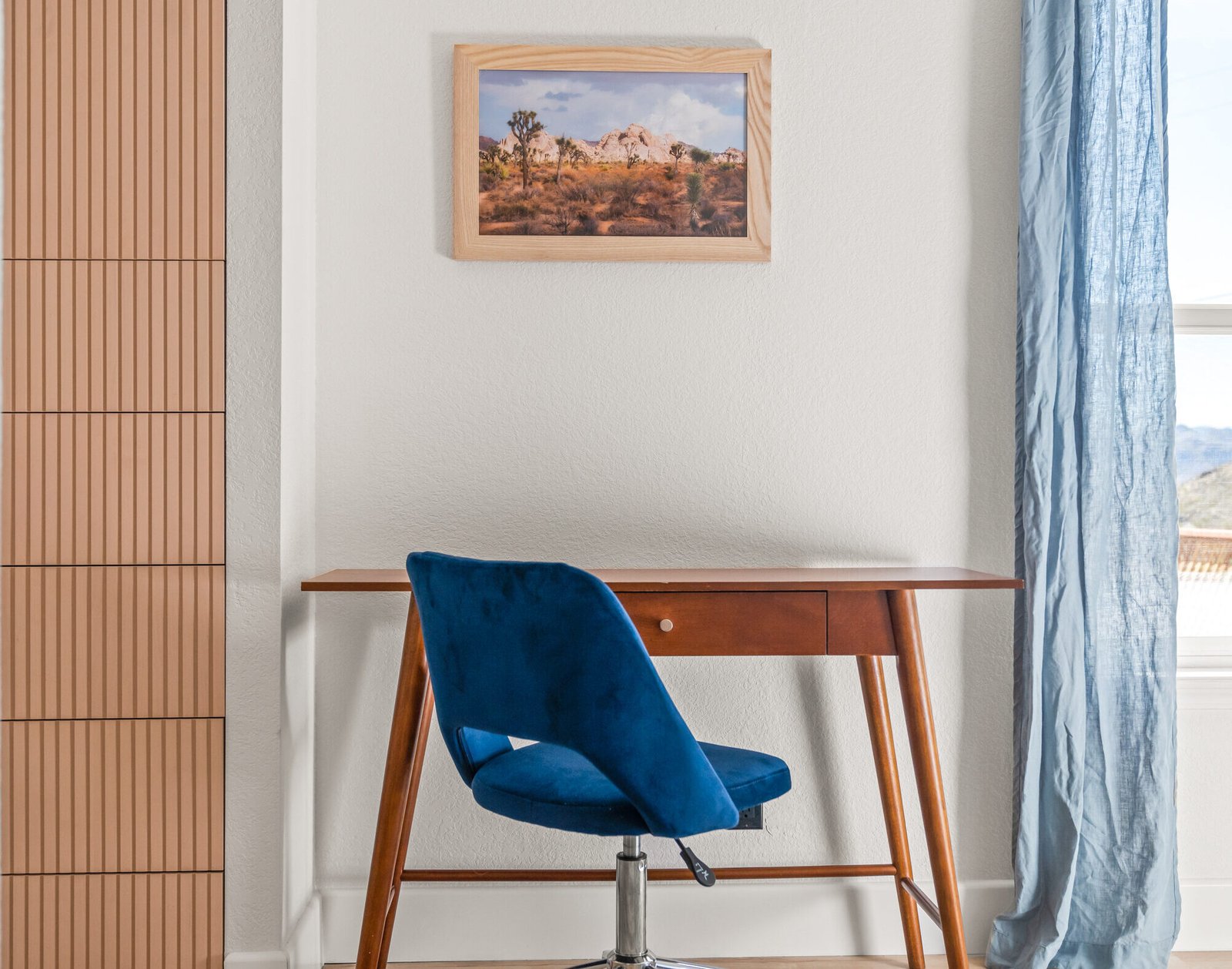 Modern home office with a wood desk, blue velvet chair, slatted wood cabinet, and framed desert photograph.